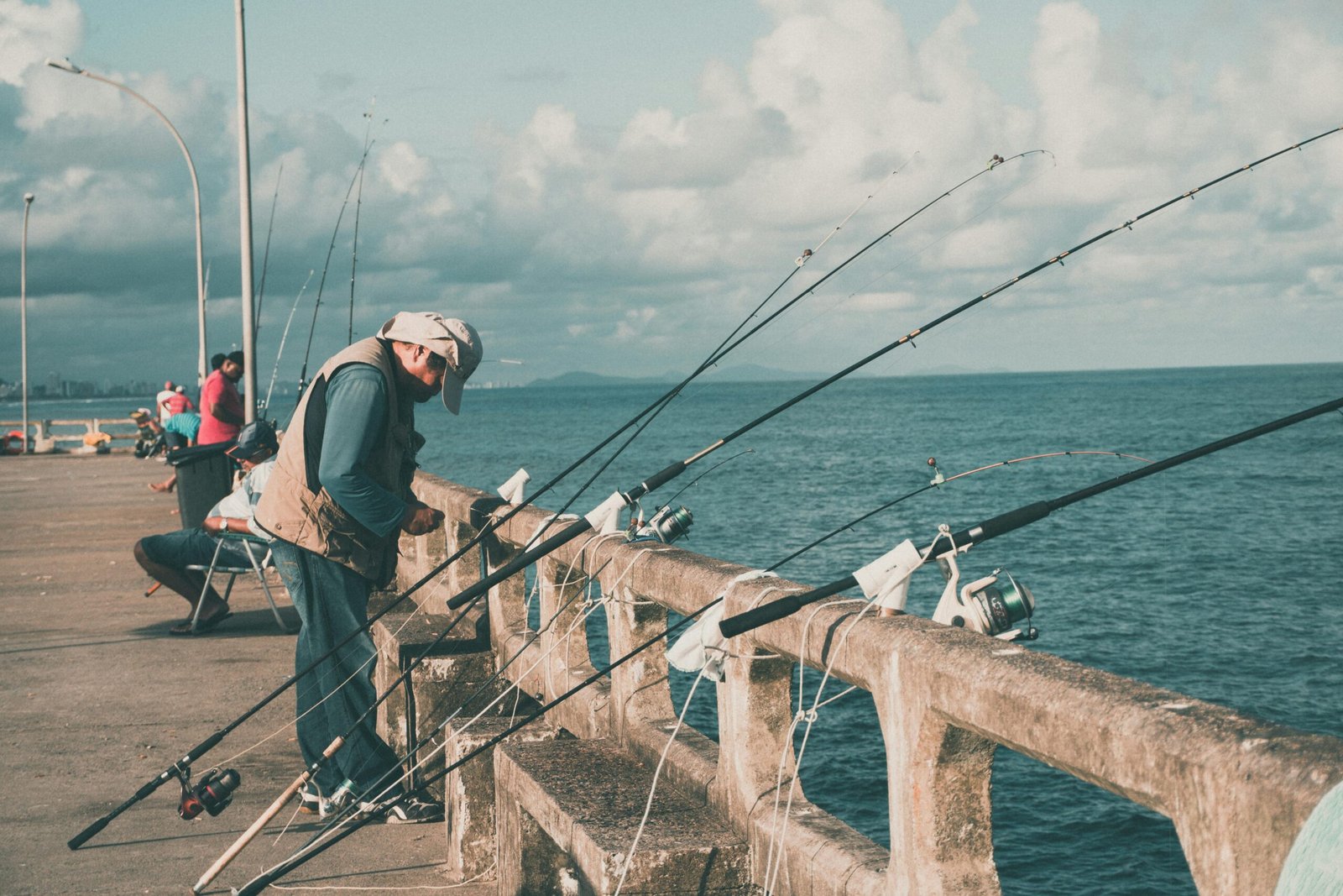 A group of people fishing from a concrete pier, with fishing rods propped along the railing and the ocean in the background under a cloudy sky.