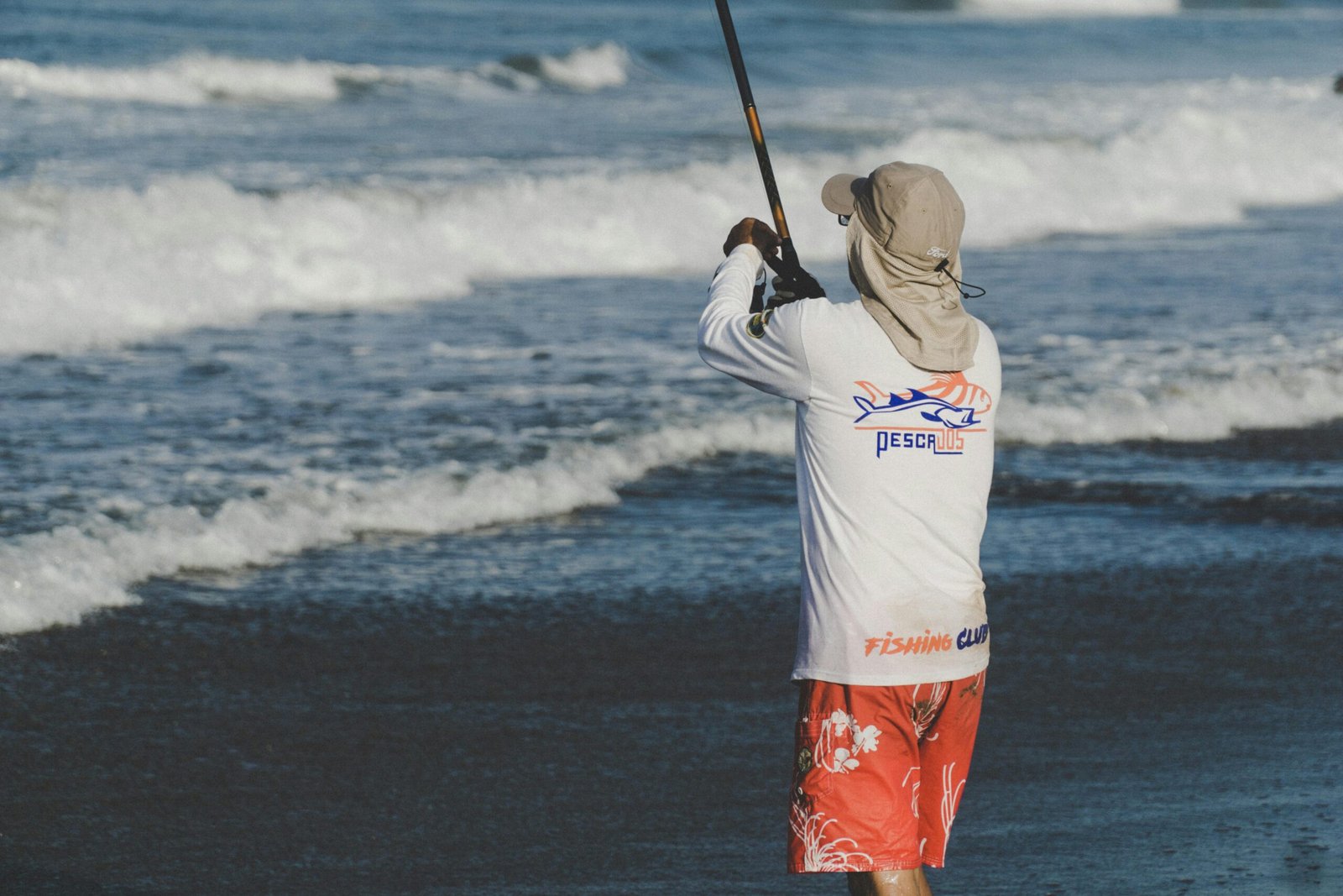 A man standing on the shore facing the ocean while casting a fishing rod, wearing a sun hat, long-sleeve fishing shirt, and red swim trunks.